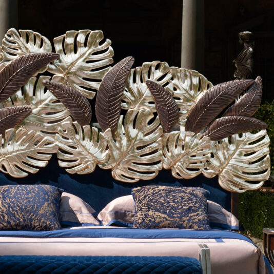 Luxurious Bed With Palm Leaf Design Headboard The Luxurious Bed With Palm Leaf Design Headboard showcases an elaborate arrangement of large metallic tropical leaves. Adorned with blue and beige bedding, complemented by blue patterned cushions, this bed is set in an elegant outdoor area surrounded by classical statues and lush greenery.