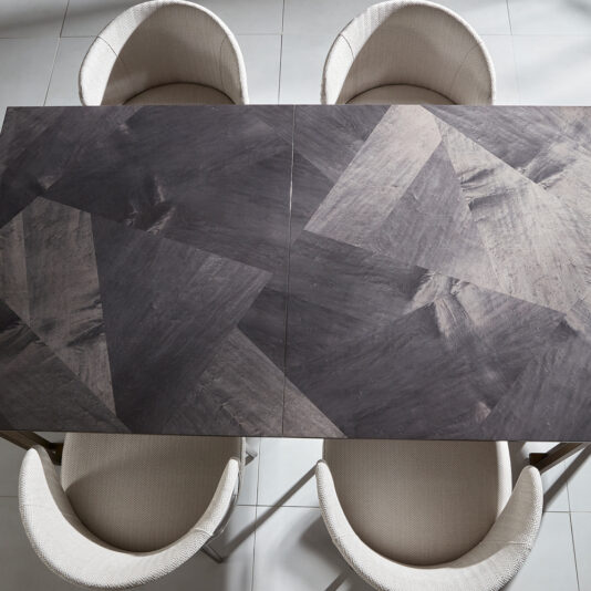 Luxury Veneer And Brass Extendable Dining Table Top-down view of the *Luxury Veneer and Brass Extendable Dining Table* featuring a geometric patterned surface in shades of gray. Four rounded, upholstered chairs in light fabric are positioned around the table, all set on a tiled floor.