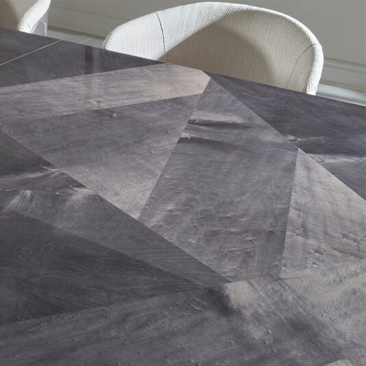 Luxury Veneer And Brass Extendable Dining Table A close-up view of the Luxury Veneer and Brass Extendable Dining Table reveals its polished, geometric patterned stone tabletop featuring various shades of gray. In the background, part of a white, textured fabric chair is visible. The reflective surface showcases intricate angles and textures that highlight the table's luxurious craftsmanship.