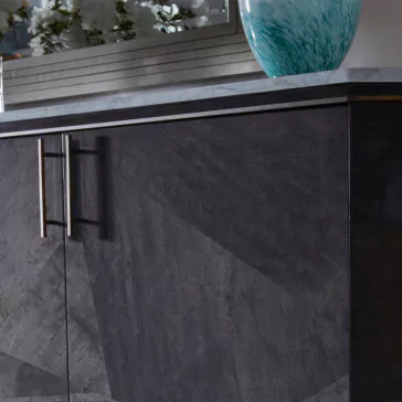 Close-up of the Luxury Marble Top Veneer Sideboard in dark gray wood, featuring stainless steel handles. A portion of its elegant marble top is visible, adorned with a turquoise decorative vase. In the reflective surface behind it, reflections of plants and a window can be seen.