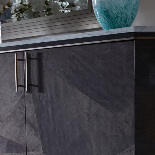 Luxury Marble Top Veneer Sideboard Close-up of the Luxury Marble Top Veneer Sideboard in dark gray wood, featuring stainless steel handles. A portion of its elegant marble top is visible, adorned with a turquoise decorative vase. In the reflective surface behind it, reflections of plants and a window can be seen.