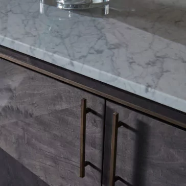 A close-up view of a kitchen countertop showcases the Luxury Marble Top Veneer Sideboard, featuring a marble top with light gray and white veining. Below, two sleek dark wood cabinet doors with vertical bronze handles and subtle wood grain patterns complete this elegant sideboard design.