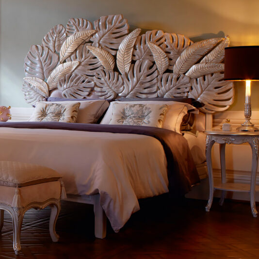 A sophisticated bedroom featuring the Luxury Bed With Tropical Palm Leaf Headboard adorned with oversized, sculpted leaves. The king-sized bed is dressed in elegant, neutral linens and flanked by ornate bedside tables. A cushioned bench sits at the foot of the bed, and the room is softly lit.
