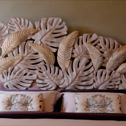 The Luxury Bed With Tropical Palm Leaf Headboard includes two intricately patterned pillows and showcases a stunning headboard adorned with numerous textured, overlapping leaf designs in various shades of beige and light brown, adding an artistic touch to the bedroom décor.