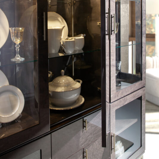 The Luxury Brown Maple Veneer Display Cabinet, with its glass doors partially open, reveals an elegant arrangement of white porcelain dishes inside. Plates, cups, and a soup tureen are neatly organized alongside a golden goblet and a silver teapot.