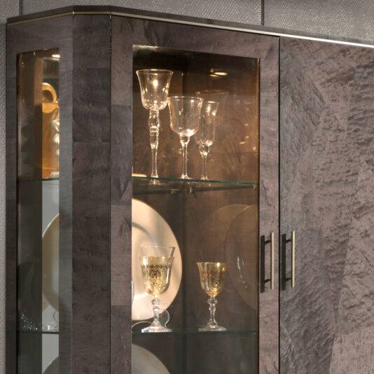 A Luxury Brown Maple Veneer Display Cabinet with glass doors, showcasing elegant glassware such as wine glasses and goblets, some adorned with gold embellishments. The shelves also feature white plates in the background.