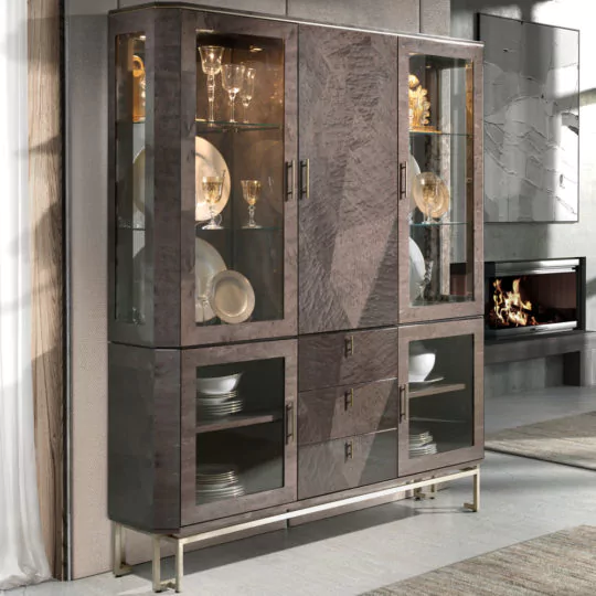 A luxury brown maple veneer display cabinet with glass doors and a modern design stands tall in a contemporary living room. The top section showcases glassware and decorative items, while the bottom section holds dinner plates and bowls. The cabinet is elevated on sleek metallic legs.