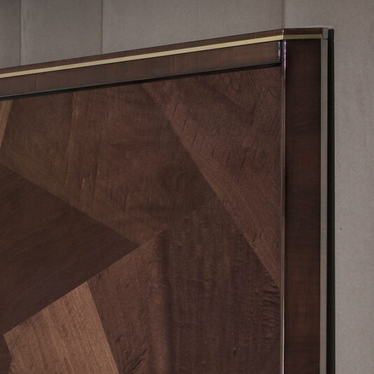 Opulent Brown Maple Veneer Bed Close-up of the Opulent Brown Maple Veneer Bed showcasing its luxurious brown maple veneer panel, designed with a geometric, angular pattern in various shades of brown. The bed features a smooth, polished surface and a border highlighted by a metallic strip along the top edge. The softly blurred background suggests an indoor setting.