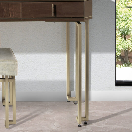 A detailed close-up of the Luxury Brown Maple Veneer Vanity Table, featuring stylish beige metal legs, set in a minimalist room. In the foreground, a coordinating beige metal bench with a light cushion is partially visible. The background showcases a window framed by greenery outside.