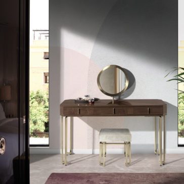 The Luxury Brown Maple Veneer Vanity Table, featuring a round mirror and several beauty products, along with a cushioned stool, sits in front of a light-colored wall. The table boasts sleek golden legs, while sunlight filters through a window, casting soft shadows. A bed and bedside lamp can be partially seen on the left side.