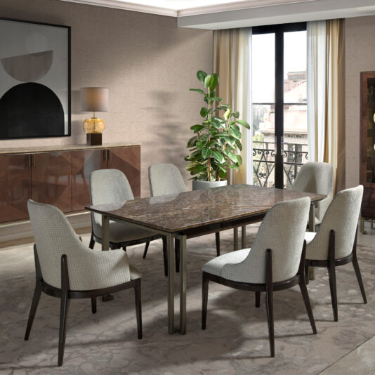 A contemporary dining room showcases a rectangular marble table complemented by the Luxury Chic Dining Set, which includes six upholstered chairs. The space is further adorned with a sideboard that holds a lamp and has artwork mounted above it. Additionally, there is a large plant in the room, and natural light filters through beige curtains covering the window that offers a balcony view. The overall decor exudes modern elegance.