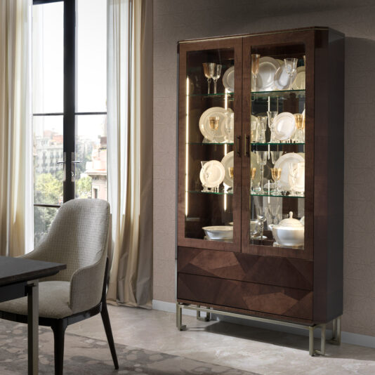 Luxury Chic Dining Set A well-lit dining room featuring the Luxury Chic Dining Set, complete with a large cabinet showcasing an assortment of glassware, dishes, and decorative items. Next to the cabinet is a cushioned dining chair and part of a stylish dining table, all framed by tall windows in the background that let in ample natural light.