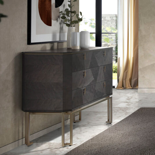 The Luxury Olive Veneer Chest Of Drawers with gold handles stands against a beige wall. This dark-wood dresser features four drawers and is elevated on slim gold legs. Atop the chest, two white vases filled with green foliage complement the abstract framed artwork above. To the right, a large window with cream curtains allows natural light to fill the room.