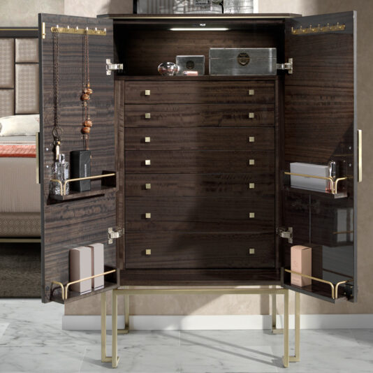 The Luxury Olive Veneer Jewellery Armoire stands elegantly against a beige wall, showcasing its dark wood interior and multiple drawers adorned with gold square handles. The inside of the doors holds an array of jewelry, perfumes, and neatly organized boxes. In the background, part of a bed is visible.