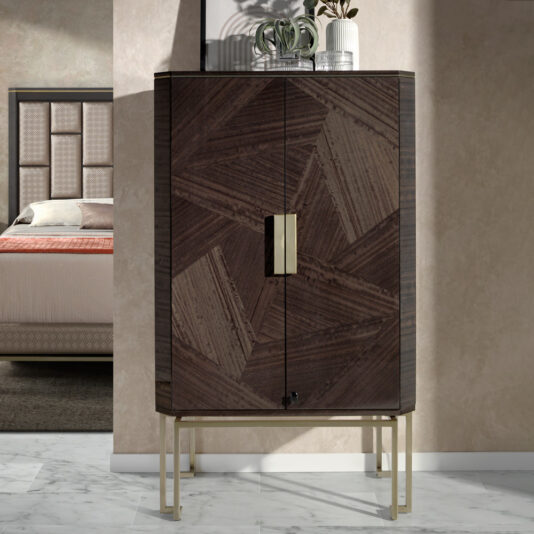 The Luxury Olive Veneer Jewellery Armoire, a modern and tall cabinet featuring geometric wooden pattern doors with gold hinges and a lock, stands on thin gold legs. Two small potted plants are placed on top of it. In the background, a bedroom with a bed and headboard is visible. The floor is marble tiled.