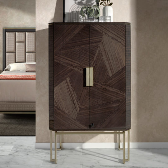 The Luxury Olive Veneer Jewellery Armoire, a modern and tall cabinet featuring geometric wooden pattern doors with gold hinges and a lock, stands on thin gold legs. Two small potted plants are placed on top of it. In the background, a bedroom with a bed and headboard is visible. The floor is marble tiled.