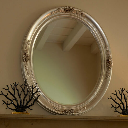 A Luxury Oval Mirror With Floral Details hangs on a beige wall above a mantel. The ornate frame is decorated with intricate floral designs. On the mantel below are two decorative black branch sculptures, one on each side of the mirror. The room’s ceiling beams are reflected in the mirror.