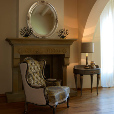 A cozy room features an ornate armchair with floral upholstery positioned in front of a stone fireplace. The mantel is adorned with the Luxury Oval Mirror With Floral Details, and beside a sunlit arched window with sheer curtains stands a side table holding a lamp and decorative items.