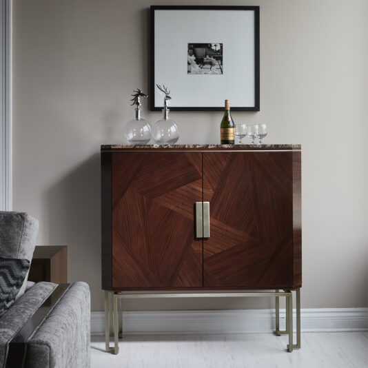 A Luxury Rosewood Veneer Drinks Cabinet with a marbled top stands against a light wall. On top, two glasses with deer-shaped handles, a bottle of wine with two wine glasses, and a framed photograph are displayed. Part of a gray sofa with a zigzag cushion is visible on the left.