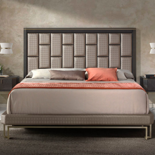 A stylish bedroom showcases the Luxury Panel Upholstered Bed, highlighted by a large, geometric-patterned headboard and adorned with an assortment of pillows in white, grey, and coral. The bed is dressed in a neutral-toned bedspread and topped with a coral throw blanket. On either side of the bed are sleek nightstands accompanied by white pendant lamps.