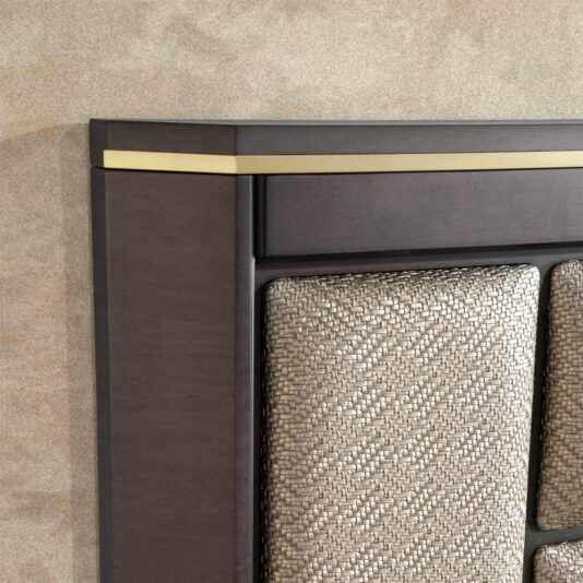 Close-up of the corner of the Luxury Panel Upholstered Bed's headboard, featuring a light woven fabric insert. The dark wooden frame is highlighted by an elegant brass detail along the top edge, set against a textured, neutral-toned wall.