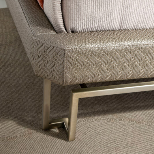 Close-up of a modern chair with textured fabric upholstery in a neutral color, reminiscent of the Luxury Panel Upholstered Bed. The chair has sleek metal legs with a metallic finish, designed in an angular, geometric shape, resting on a beige carpet. The upholstery fabric features a subtle, woven pattern.
