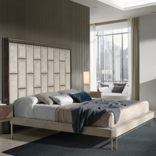 Modern bedroom with a Luxury Velvet Fabric Upholstered Bed showcasing a beige tufted headboard. The bed is dressed in gray and white bedding, complemented by a dark blue throw blanket. On either side of the bed are two bedside tables, each adorned with lamps. A large window in the background frames a view of trees and bathes the room in natural light.