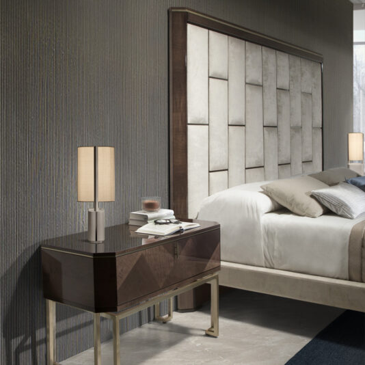 Luxury Velvet Fabric Upholstered Bed A modern bedroom showcases the Luxury Velvet Fabric Upholstered Bed, complete with a large, padded headboard. Beside the bed stands a sleek, dark wooden nightstand adorned with a stylish lamp, a book, glasses, and a cup. The walls and flooring are in neutral tones.