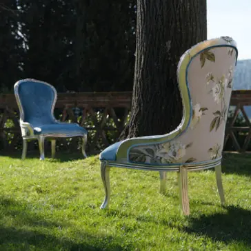 Luxury Venetian Style Occasional Chair Two Luxury Venetian Style Occasional Chairs are placed on a grassy lawn beneath a large tree. One chair is upholstered in blue fabric, while the other features a floral pattern. A brick railing in the background overlooks a scenic, distant hilly landscape.
