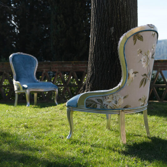 Two Luxury Venetian Style Occasional Chairs are placed on a grassy lawn beneath a large tree. One chair is upholstered in blue fabric, while the other features a floral pattern. A brick railing in the background overlooks a scenic, distant hilly landscape.
