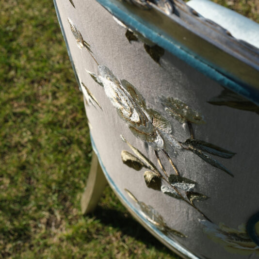 A detailed close-up of the back of a Luxury Venetian Style Occasional Chair highlights its embroidered floral pattern, showcasing intricate green and golden leaves and petals. The chair is elegantly situated outdoors on a lush patch of green grass.