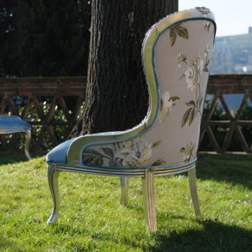 Luxury Venetian Style Occasional Chair A Luxury Venetian Style Occasional Chair with floral upholstery and blue accents is placed on a grassy lawn near a tree. In the background, there's a brick fence and a scenic view of a hilly landscape under a clear sky. Another such Luxury Venetian Style Occasional Chair is partially visible to the left.