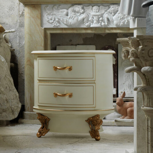 The Luxury Venetian Style Oval Bedside Cabinet is an elegant, luxury piece designed with two drawers featuring ornate gold handles and decorative gold accents on the base. It is set in a richly decorated, classical space with carved stone and marble elements in the background.