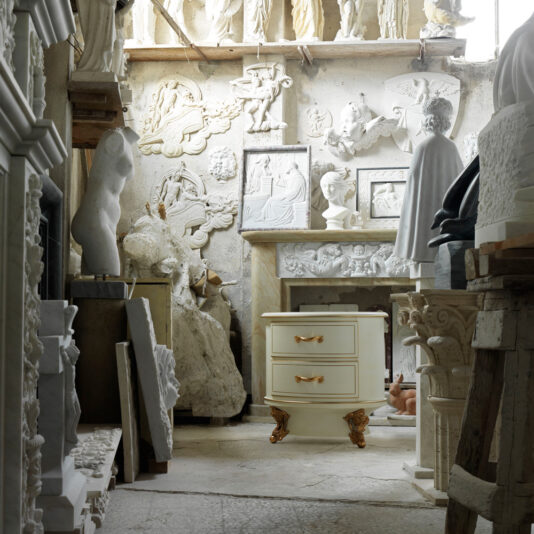 Luxury Venetian Style Oval Bedside Cabinet A cluttered workshop filled with classical-style sculptures and wall reliefs houses a variety of statues, busts, and decorative elements in white and beige tones. At the center stands the Luxury Venetian Style Oval Bedside Cabinet, an ornate piece adorned with gold handles.