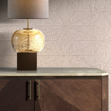 A table lamp with a textured beige shade and a round, speckled glass base sits atop the Marble Top Brown Maple Sideboard, which boasts modern geometric detailing and metallic handles. The backdrop highlights a wall adorned with a geometric pattern.