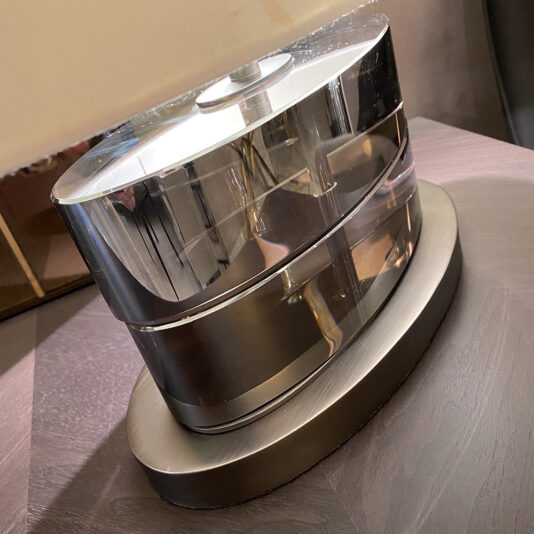 A Mid Century Smoked Crystal Table Lamp, characterized by its sleek design and cylindrical central element, features an oval-shaped clear base with a metallic finish. This contemporary piece sits elegantly on a wooden surface.