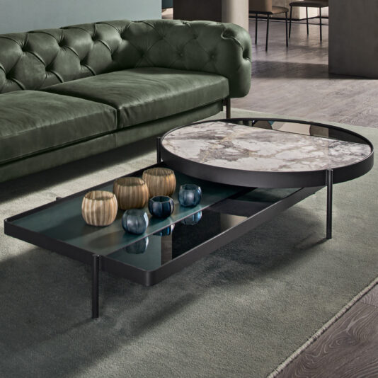Modern living room with a green tufted sofa and the "Modern Glass And Marble Modular Coffee Table." The table features a marble top and a glass lower shelf, holding various decorative items including gold and blue candle holders. The room has a muted color palette with soft lighting.