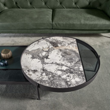 The Modern Glass And Marble Modular Coffee Table, boasting a distinctive split surface design with natural gray marble on one side and dark-tinted glass on the other, stands elegantly on thin metal legs. It rests on a plush gray carpet in front of a green tufted sofa, with decorative items neatly arranged on its lower shelf.