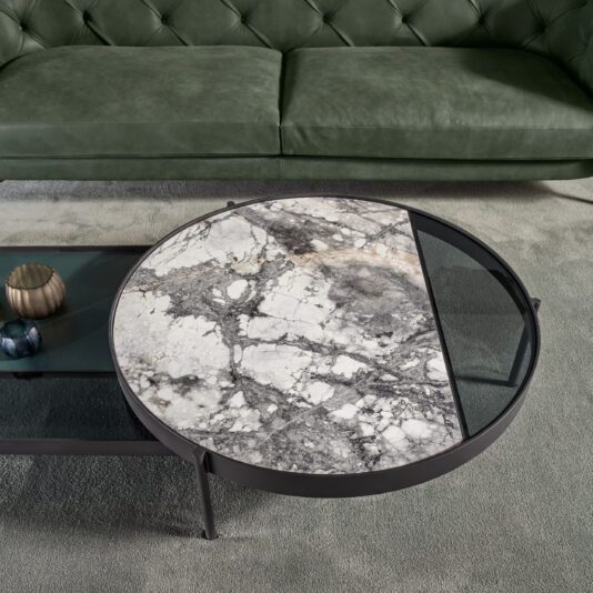 Modern Glass And Marble Modular Coffee Table The Modern Glass And Marble Modular Coffee Table, boasting a distinctive split surface design with natural gray marble on one side and dark-tinted glass on the other, stands elegantly on thin metal legs. It rests on a plush gray carpet in front of a green tufted sofa, with decorative items neatly arranged on its lower shelf.