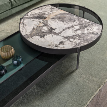 The Modern Glass and Marble Modular Coffee Table boasts a contemporary round design with a sleek black metal frame. Its unique tabletop is divided into two sections; one showcases a luxurious marble surface, while the other features stylish black transparent glass. This elegant piece is complemented by its placement on a green textured rug and adorned with decorative glass items.