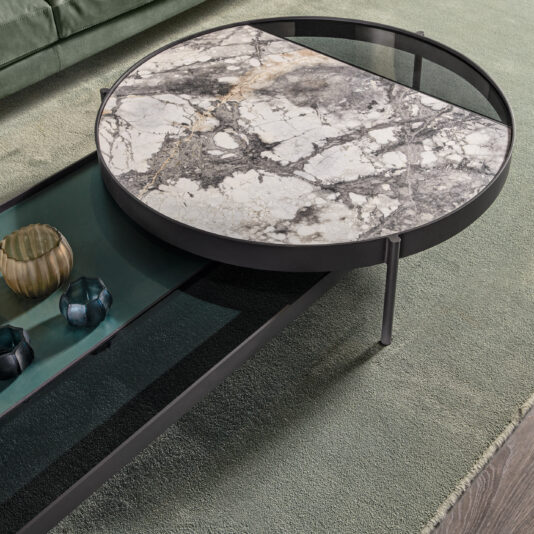 Modern Glass And Marble Modular Coffee Table The Modern Glass and Marble Modular Coffee Table boasts a contemporary round design with a sleek black metal frame. Its unique tabletop is divided into two sections; one showcases a luxurious marble surface, while the other features stylish black transparent glass. This elegant piece is complemented by its placement on a green textured rug and adorned with decorative glass items.