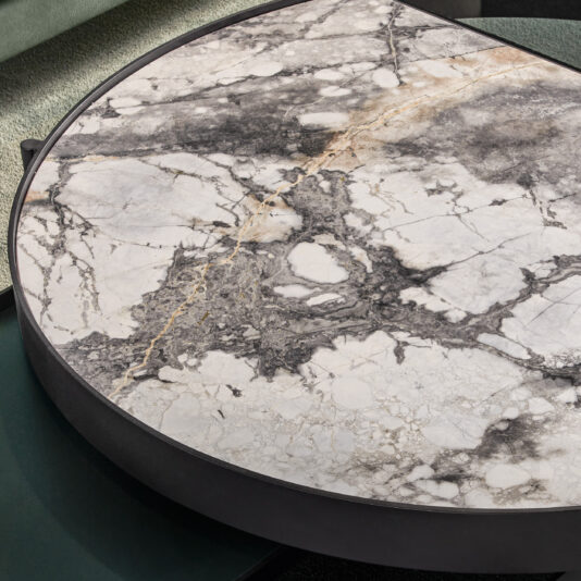 Modern Glass And Marble Modular Coffee Table A close-up view of the Modern Glass And Marble Modular Coffee Table highlights its marbled surface with intricate natural vein patterns in shades of white, grey, and black. The table boasts a dark circular rim and is set against a green, textured background.