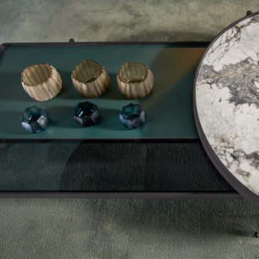 Here's a top-down view of the Modern Glass And Marble Modular Coffee Table. This setup showcases a circular section with a marble-like texture and an adjacent rectangular section. The glass surface is adorned with four decorative candle holders, arranged neatly: two beige and two blue. The entire table sits elegantly on a green carpet.