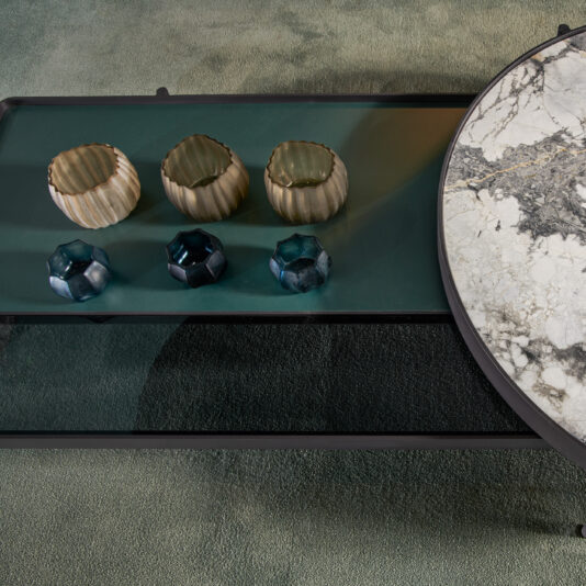 Modern Glass And Marble Modular Coffee Table Here's a top-down view of the Modern Glass And Marble Modular Coffee Table. This setup showcases a circular section with a marble-like texture and an adjacent rectangular section. The glass surface is adorned with four decorative candle holders, arranged neatly: two beige and two blue. The entire table sits elegantly on a green carpet.
