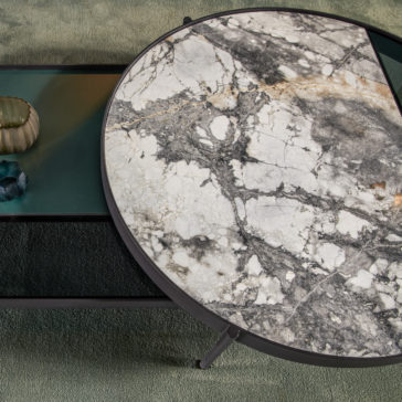 The Modern Glass And Marble Modular Coffee Table showcases a large round segment with a cracked marble top and an opaque black-finished rectangular glass section. A textured green object adorns the table, which is placed on a soft green carpet.