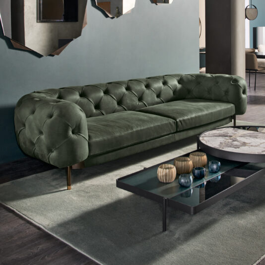 A stylish living room features the Modern Low Button Upholstered Suede Sofa in plush green with rounded arms, positioned against a dark green wall. In front of the sofa is a sleek coffee table with a marble top and several decorative items on it, all placed on a grey rug over a dark wood floor.