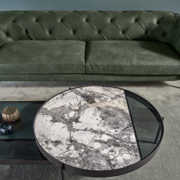 Modern Low Button Upholstered Suede Sofa A living room featuring a modern low button upholstered suede sofa in green and a round marble coffee table with a metal frame. The table has a geometric design with sections of marble and glass. A small gold candle holder and blue decorative items are on the table, enhancing the contemporary ambiance.