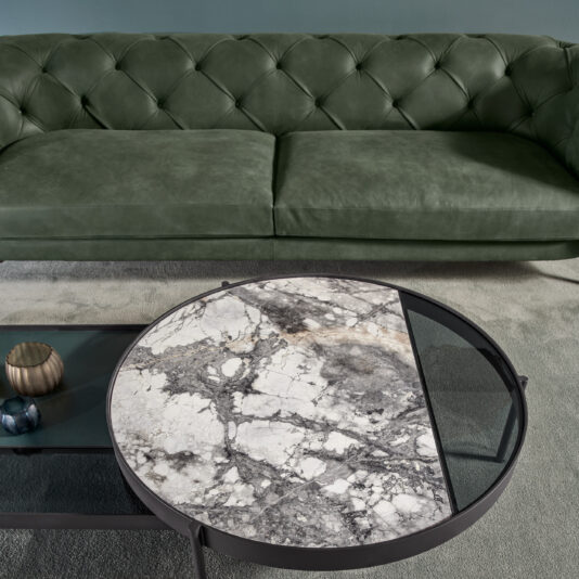 A living room featuring a modern low button upholstered suede sofa in green and a round marble coffee table with a metal frame. The table has a geometric design with sections of marble and glass. A small gold candle holder and blue decorative items are on the table, enhancing the contemporary ambiance.