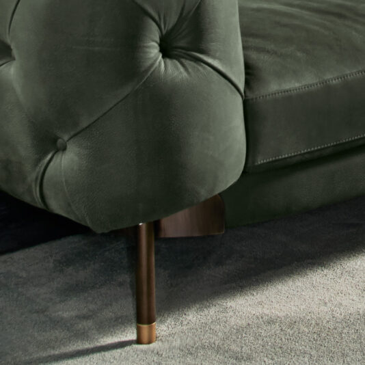 Close-up of the armrest of a Modern Low Button Upholstered Suede Sofa in dark green, featuring visible button-tuft detailing and wooden legs. The sofa is set against a plush grey carpet, highlighting the soft textures of both the suede fabric and the carpet.