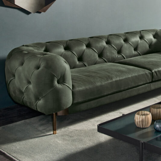 A Modern Low Button Upholstered Suede Sofa in dark green velvet with a tufted back and armrests sits against a muted gray wall. A black coffee table in front of the sofa holds three decorative wooden bowls. The room's lighting casts soft shadows, adding to the cozy atmosphere.
